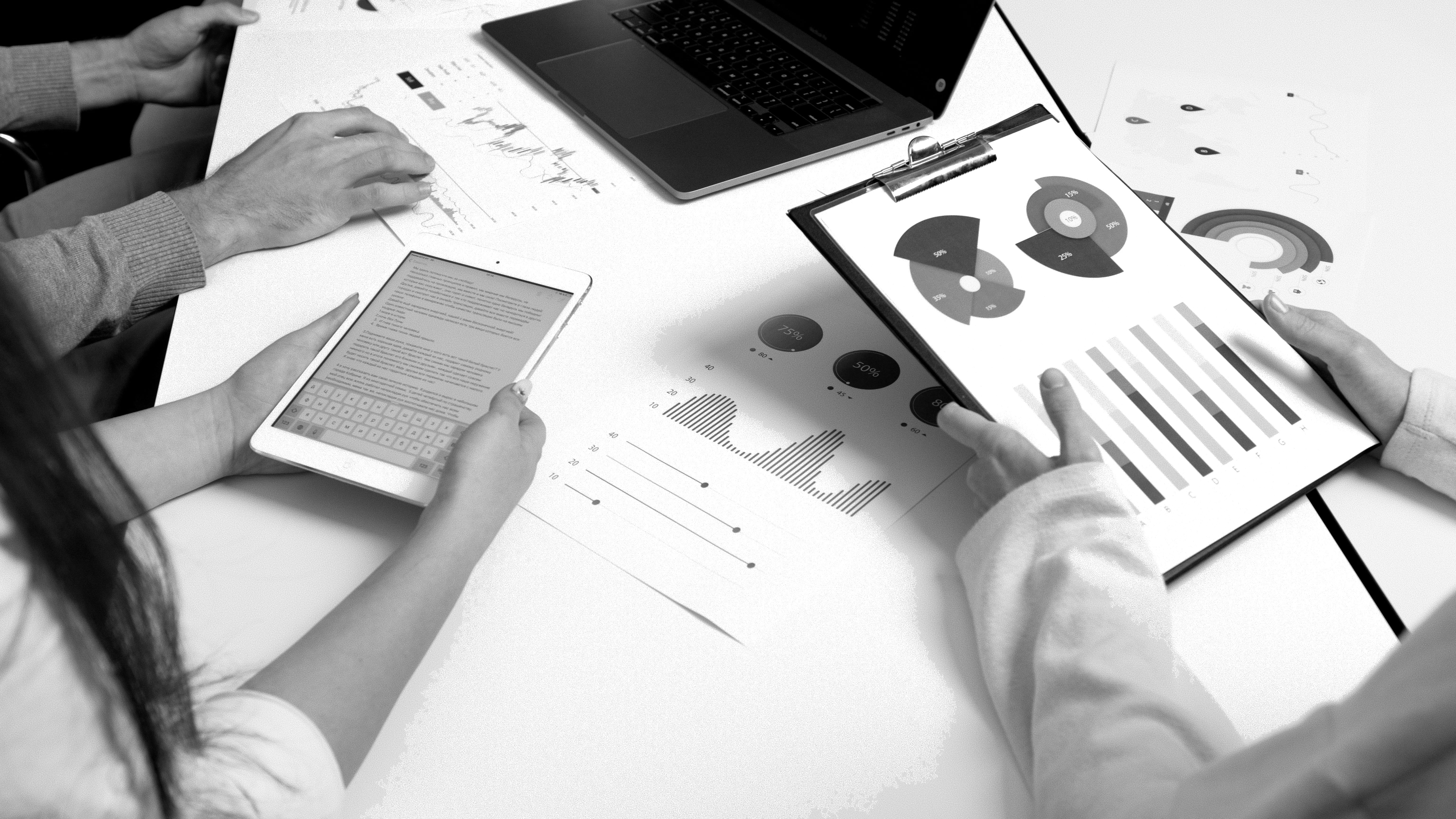 black and white photo of laptop, marketing book, headphones, and person's hands writing on notepad